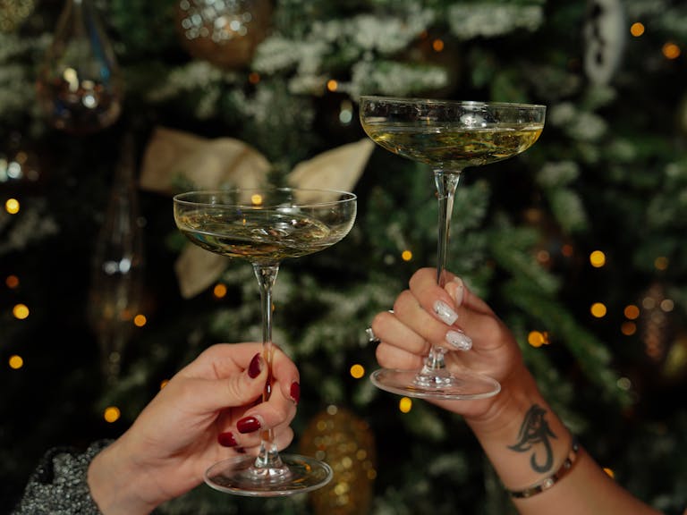 Two women clinking champagne glasses in front of a decorated Christmas tree, capturing a festive holiday moment.