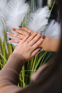 Close-up of a woman's hands adorned with elegant jewelry and stylish nail art against a decorative feather background.