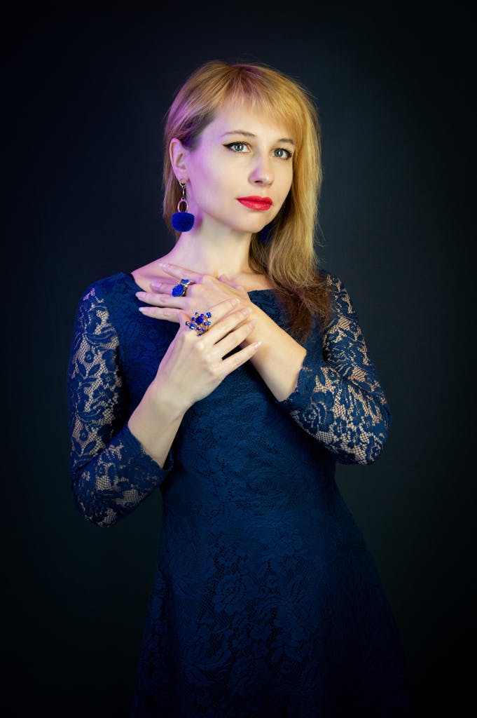 Portrait of a woman in a blue lace dress with jewelry, posing against a dark background.