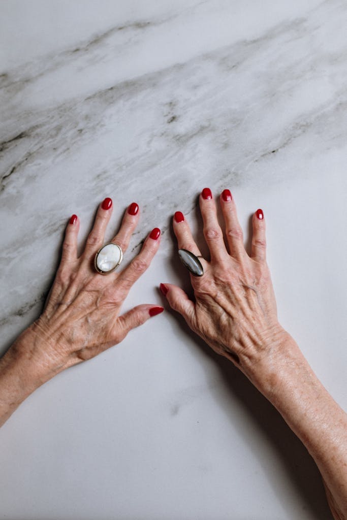 An elderly woman's hands with red nail polish and jewelry on a marble surface.