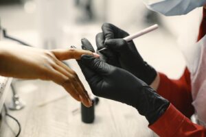 Manikiurzystka w czarnych rękawiczkach przygotowuje płytkę paznokci klientki przed nałożeniem primera i bazy