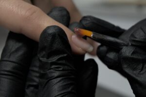 Dłonie w czarnych rękawiczkach nakładają pędzelkiem żel na naturalny paznokieć podczas manicure